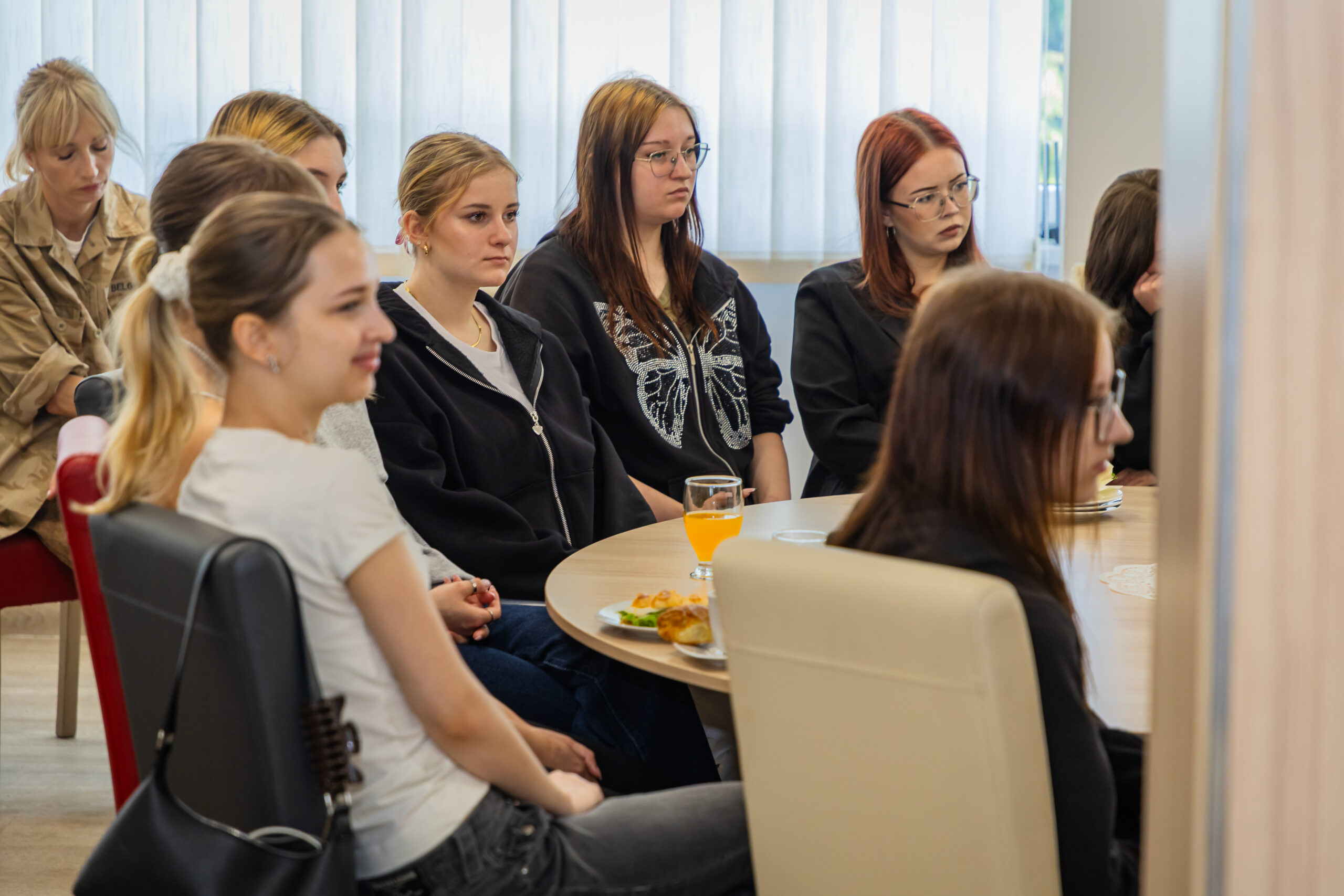 Grupa młodych kobiet siedzi blisko siebie przy stole, uważnie słuchając. Niektóre noszą czarne lub białe ubrania. Na stole znajduje się talerz z jedzeniem i szklanka soku pomarańczowego. Okno z białymi żaluzjami wpuszcza naturalne światło z tyłu.