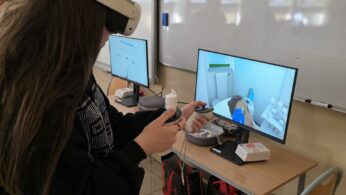 Osoba nosząca gogle VR i trzymająca kontrolery wchodzi w interakcję z symulacją medyczną wyświetlaną na monitorze w klasie. Na biurku znajdują się akcesoria VR i pudełka, a dwa ekrany wyświetlają powiązane wizualizacje. W klasie znajduje się tablica i podłoga wyłożona kafelkami.