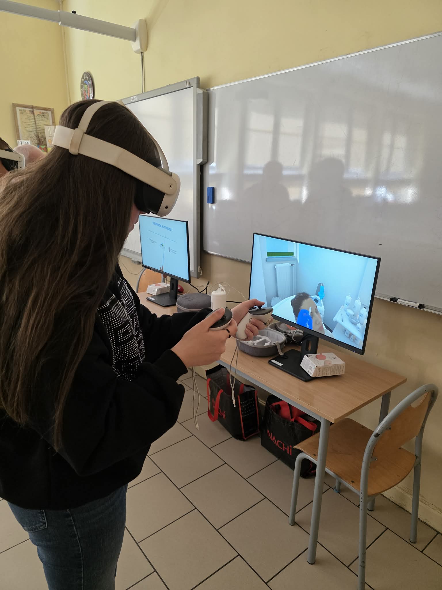 Osoba nosząca gogle VR i trzymająca kontrolery wchodzi w interakcję z symulacją medyczną wyświetlaną na monitorze w klasie. Na biurku znajdują się akcesoria VR i pudełka, a dwa ekrany wyświetlają powiązane wizualizacje. W klasie znajduje się tablica i podłoga wyłożona kafelkami.
