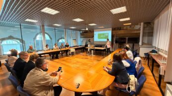 Wnętrze dużej sali konferencyjnej z drewnianym stołem w kształcie litery U, przy którym siedzą uczestnicy spotkania. Na końcu sali, przy ekranie projekcyjnym wyświetlającym prezentację, stoją dwie prelegentki. Pomieszczenie ma kasetonowy sufit i duże przeszklenia po lewej stronie.