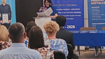 Kobieta stoi na podium, przemawiając do publiczności w klasie lub sali konferencyjnej. Trzyma teczkę i uśmiecha się. Za nią znajdują się niebieskie banery z polskim tekstem, zegar i oprawione polskie godło na ścianie. Ludzie siedzą naprzeciwko niej, słuchając uważnie.