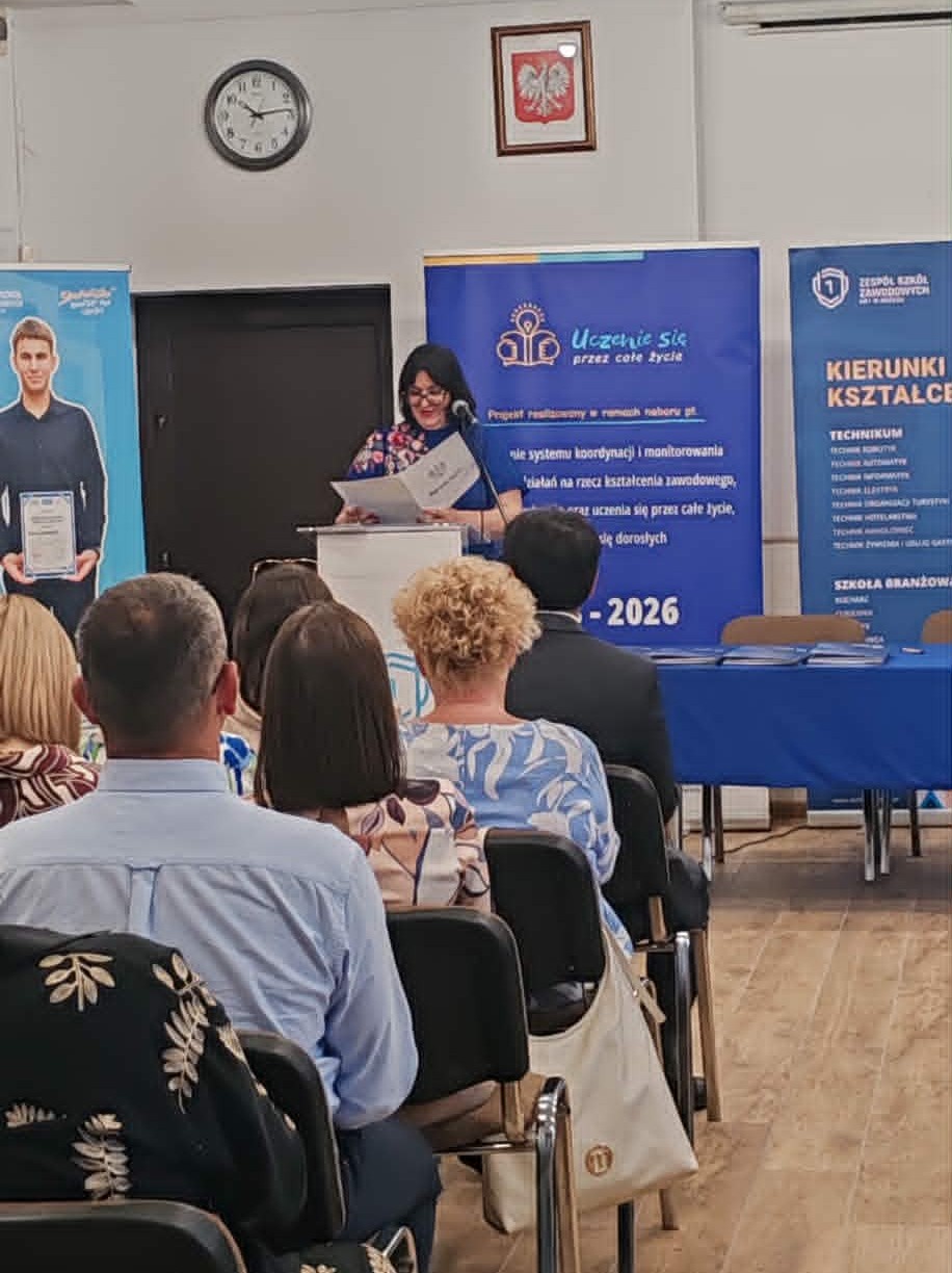 Kobieta stoi na podium, przemawiając do publiczności w klasie lub sali konferencyjnej. Trzyma teczkę i uśmiecha się. Za nią znajdują się niebieskie banery z polskim tekstem, zegar i oprawione polskie godło na ścianie. Ludzie siedzą naprzeciwko niej, słuchając uważnie.
