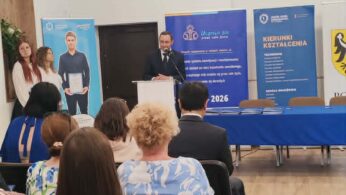 Mężczyzna w garniturze przemawia na podium w jasnym pomieszczeniu z drewnianą podłogą, twarzą do publiczności. Za nim znajdują się niebieskie banery informacyjne i stół z niebieskim obrusem. Dwie kobiety stoją z boku. Publiczność siedzi i uważnie słucha.