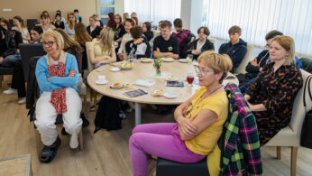 Grupa ludzi, zarówno dorosłych, jak i nastolatków, siedzi uważnie przy stołach w jasnym pomieszczeniu z dużymi oknami. Niektórzy mają przed sobą napoje i ciastka. Większość jest zwrócona do przodu, wyglądając na zaangażowanych w prezentację lub rozmowę.
