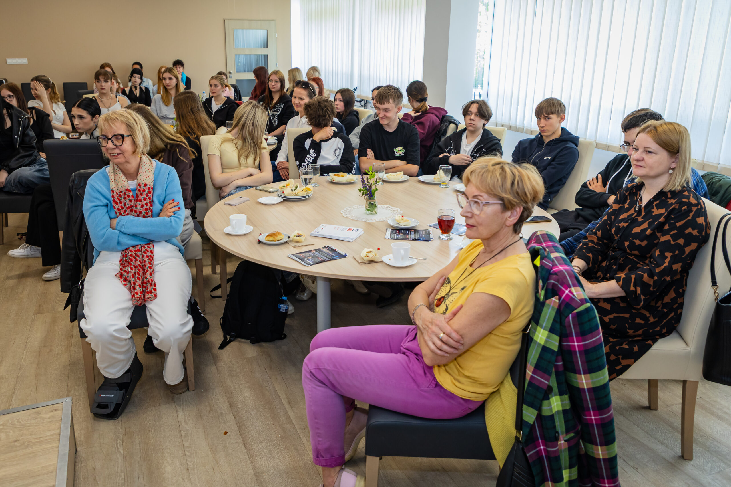 Grupa ludzi, zarówno dorosłych, jak i nastolatków, siedzi uważnie przy stołach w jasnym pomieszczeniu z dużymi oknami. Niektórzy mają przed sobą napoje i ciastka. Większość jest zwrócona do przodu, wyglądając na zaangażowanych w prezentację lub rozmowę.