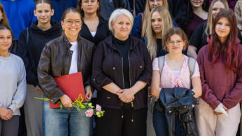 Grupa ludzi pozuje na zewnątrz, większość z nich uśmiecha się do kamery. Z przodu trzy kobiety stoją blisko siebie; jedna trzyma kwiaty i czerwoną teczkę. Inni stoją za nimi, ubrani w zwykłe ubrania. Tło jest nieostre, co sugeruje wydarzenie szkolne lub społeczne.