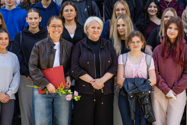 Grupa ludzi pozuje na zewnątrz, większość z nich uśmiecha się do kamery. Z przodu trzy kobiety stoją blisko siebie; jedna trzyma kwiaty i czerwoną teczkę. Inni stoją za nimi, ubrani w zwykłe ubrania. Tło jest nieostre, co sugeruje wydarzenie szkolne lub społeczne.