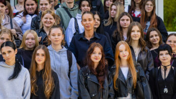 Duża grupa studentów, głównie młodych kobiet, stoi razem na zewnątrz, aby zrobić zdjęcie grupowe. Większość z nich ma na sobie swobodne ubrania, takie jak bluzy z kapturem i skórzane kurtki. Nastrój wydaje się pozytywny i przyjazny, z mieszanką uśmiechów i neutralnych wyrazów twarzy. Za nimi widoczne są drzewa.
