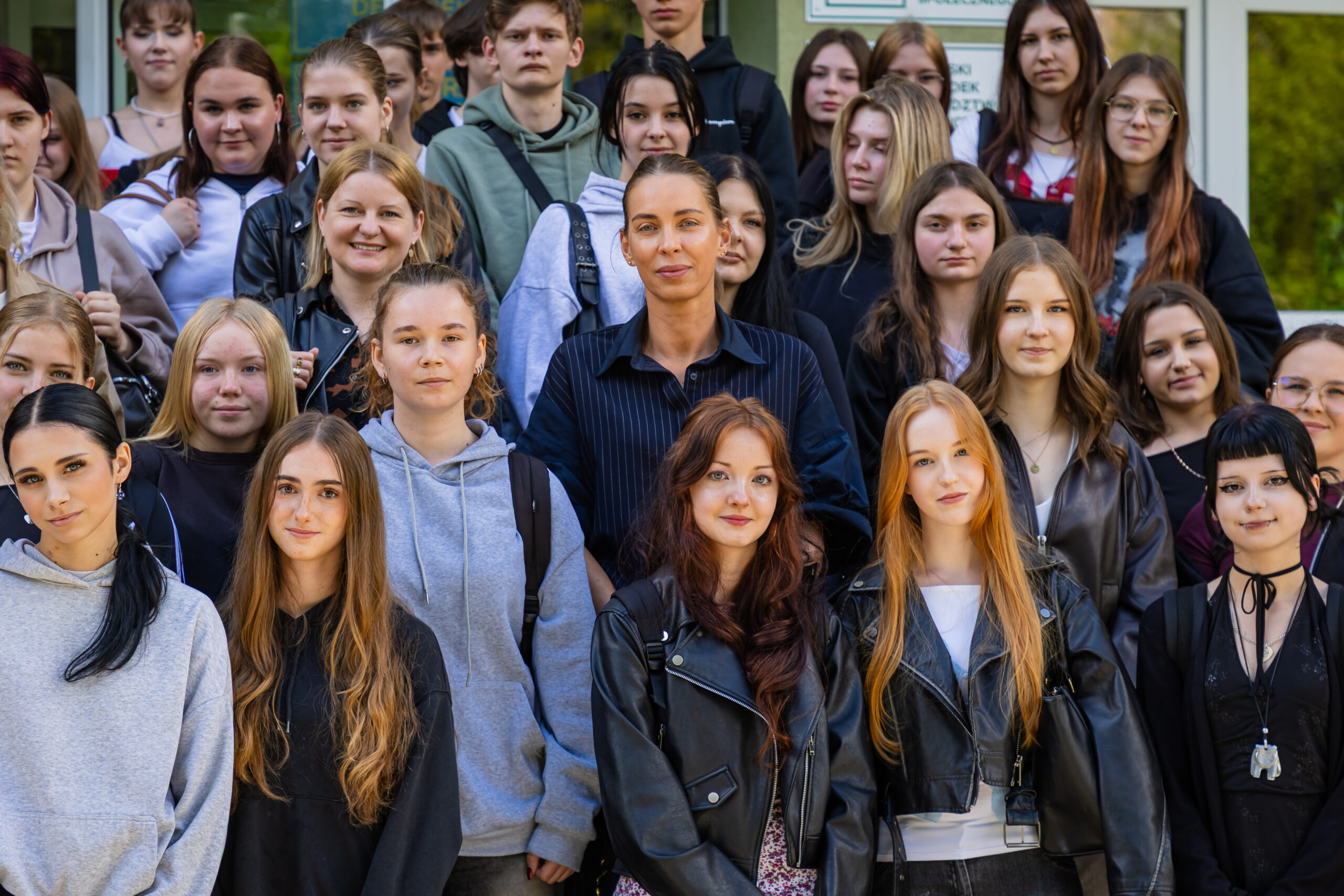 Duża grupa studentów, głównie młodych kobiet, stoi razem na zewnątrz, aby zrobić zdjęcie grupowe. Większość z nich ma na sobie swobodne ubrania, takie jak bluzy z kapturem i skórzane kurtki. Nastrój wydaje się pozytywny i przyjazny, z mieszanką uśmiechów i neutralnych wyrazów twarzy. Za nimi widoczne są drzewa.