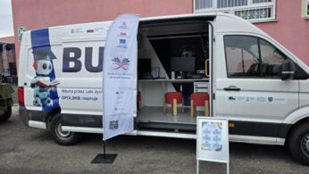 Widok z zewnątrz na biały furgon pełniący funkcję mobilnego centrum edukacyjnego, zaparkowany przed budynkiem. Na karoserii widnieje grafika z robotem w birecie oraz hasło „Nauka przez całe życie – OPOLSKIE inspiruje”. Drzwi przesuwne są otwarte, ukazując wewnątrz nowoczesne stanowiska komputerowe z czerwonymi pufami. Przed pojazdem stoi biała flaga reklamowa oraz tablica informacyjna.