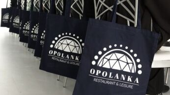Mężczyzna w białej bluzie kucharskiej i wysokiej czapce, stojący w centrum, obsługuje panel sterowania maszyny przemysłowej z rolką folii. Po prawej stronie stoi grupa młodzieży w foliowych czepkach i fartuchach ochronnych, uważnie obserwująca jego działania. Tło stanowi jasne, sterylne pomieszczenie z metalowymi stołami i sprzętem kuchennym. Jeden z chłopców w grupie ma skrzyżowane ręce na piersi.