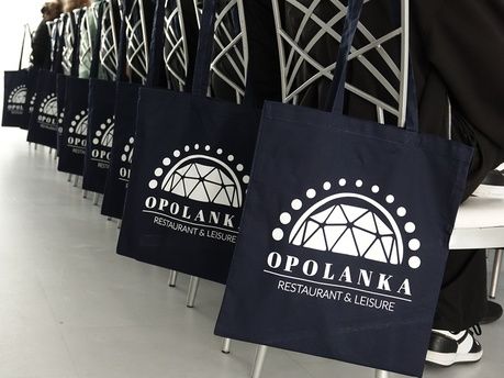 Mężczyzna w białej bluzie kucharskiej i wysokiej czapce, stojący w centrum, obsługuje panel sterowania maszyny przemysłowej z rolką folii. Po prawej stronie stoi grupa młodzieży w foliowych czepkach i fartuchach ochronnych, uważnie obserwująca jego działania. Tło stanowi jasne, sterylne pomieszczenie z metalowymi stołami i sprzętem kuchennym. Jeden z chłopców w grupie ma skrzyżowane ręce na piersi.