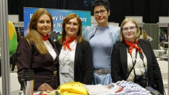 Cztery kobiety stoją rzędem za białym stołem konferencyjnym, uśmiechając się do aparatu. Trzy z nich mają na szyi identyczne czerwone apaszki, a jedna na pierwszym planie po prawej ma aparat fotograficzny przewieszony przez ramię. Na stole leżą materiały promocyjne, smycze, koszulki i długopisy. Tło jest ciemne i niewyraźne, sugerując wnętrze hali lub biura.