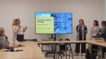 Spotkanie biznesowe lub konferencja w jasnej sali, gdzie grupa osób siedzi przy dużym, jasnym stole konferencyjnym. Po lewej stronie kobieta na stojąco prowadzi prezentację, gestykulując dłońmi. W centrum uwagi znajduje się duży ekran telewizyjny z napisem „Visita de estudo a Portugal Delegação Polaca”.