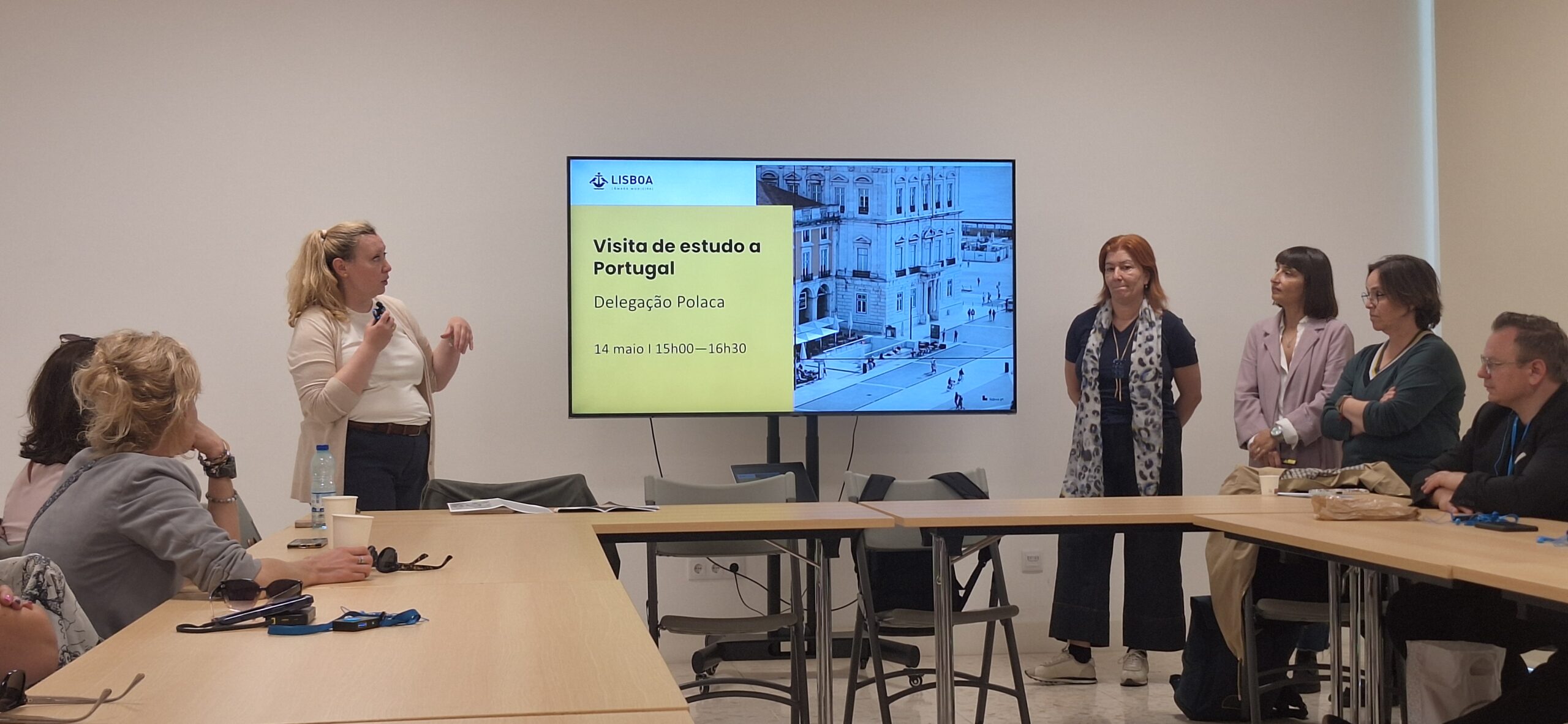 Spotkanie biznesowe lub konferencja w jasnej sali, gdzie grupa osób siedzi przy dużym, jasnym stole konferencyjnym. Po lewej stronie kobieta na stojąco prowadzi prezentację, gestykulując dłońmi. W centrum uwagi znajduje się duży ekran telewizyjny z napisem „Visita de estudo a Portugal Delegação Polaca”.