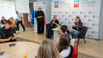 Grupa osób uczestniczy w panelu dyskusyjnym w nowoczesnej sali konferencyjnej. Trzy kobiety przemawiają z przodu; dwie siedzą z mikrofonami, a jedna stoi. Uczestnicy siedzą przy okrągłych stołach z napojami. Za prelegentami znajdują się banery z logo 