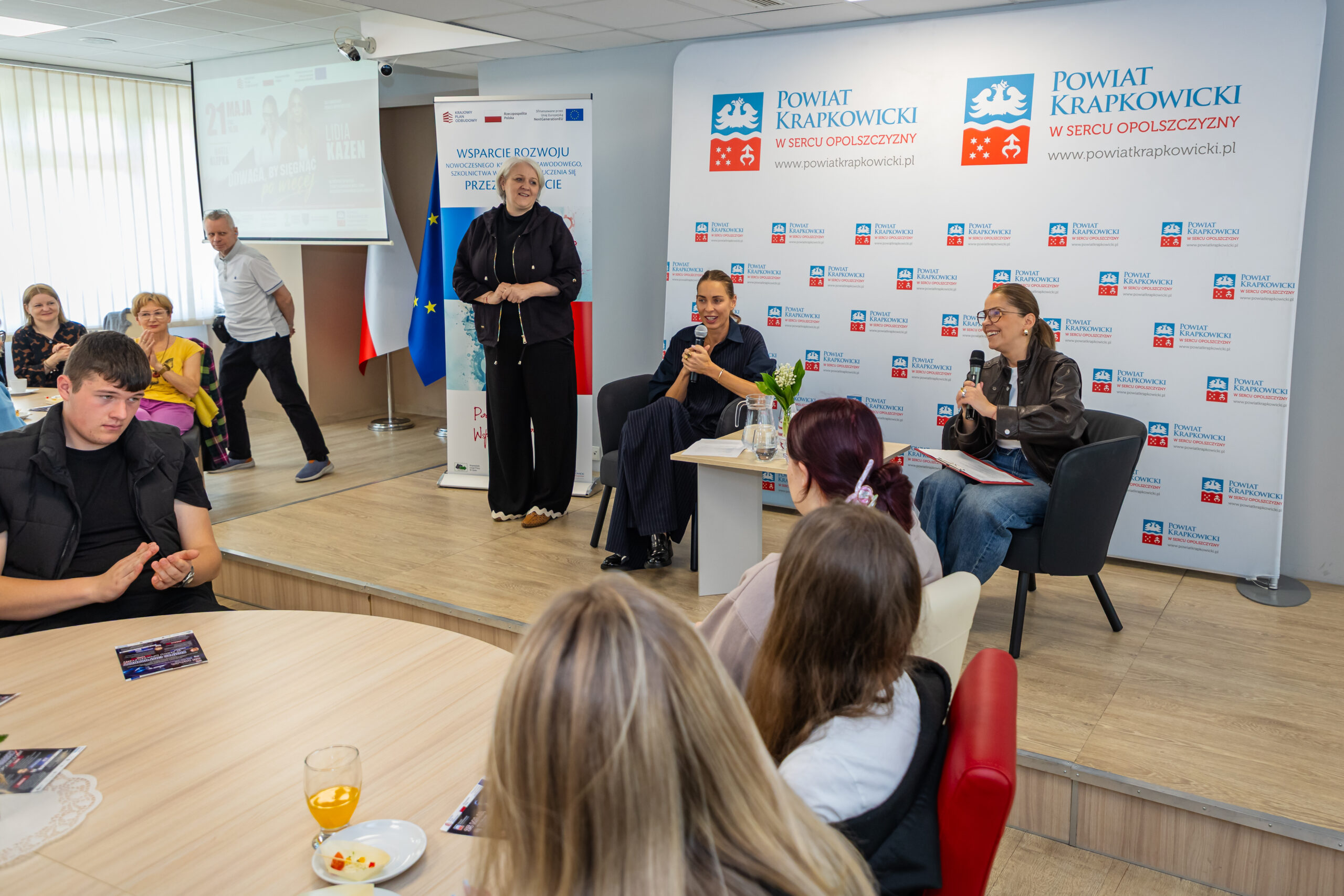 Grupa osób uczestniczy w panelu dyskusyjnym w nowoczesnej sali konferencyjnej. Trzy kobiety przemawiają z przodu; dwie siedzą z mikrofonami, a jedna stoi. Uczestnicy siedzą przy okrągłych stołach z napojami. Za prelegentami znajdują się banery z logo 