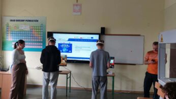 Cztery osoby stoją w klasie ze sprzętem naukowym. Dwie osoby w środku noszą zestawy VR i używają tabletów. Duży ekran wyświetla stronę internetową. Na ścianie wisi układ okresowy, a jedna osoba patrzy na smartfon po prawej stronie.
