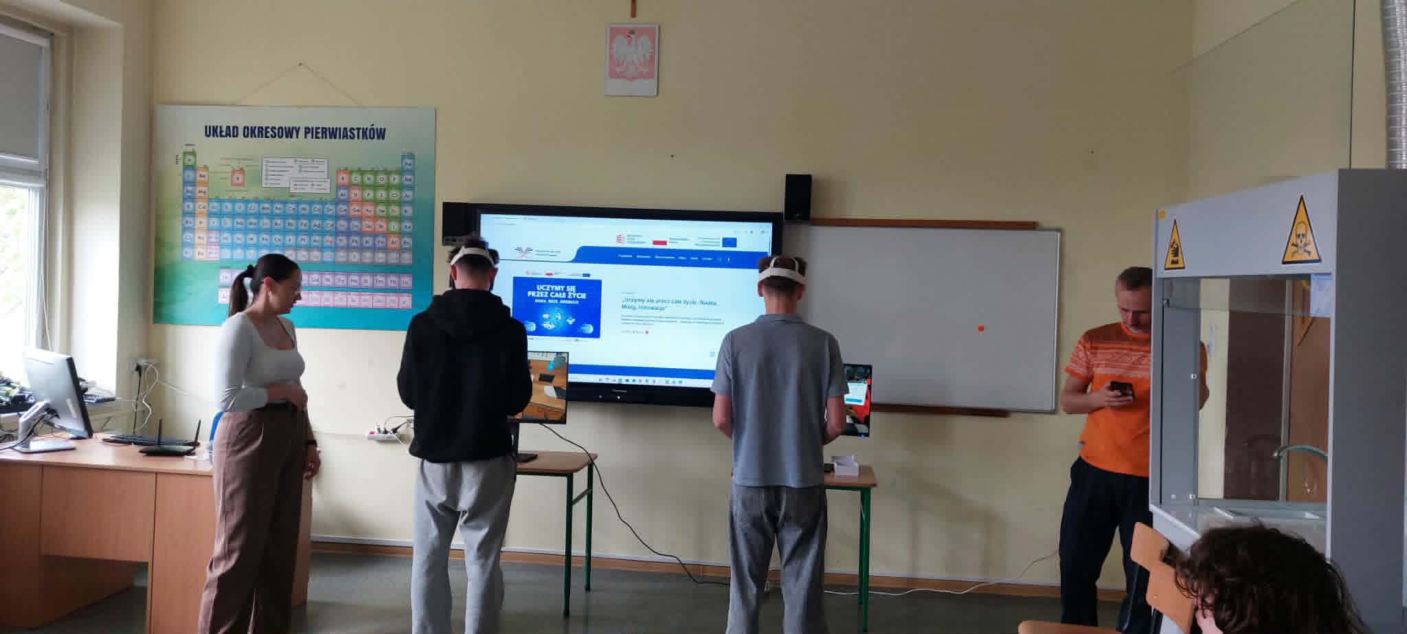 Cztery osoby stoją w klasie ze sprzętem naukowym. Dwie osoby w środku noszą zestawy VR i używają tabletów. Duży ekran wyświetla stronę internetową. Na ścianie wisi układ okresowy, a jedna osoba patrzy na smartfon po prawej stronie.