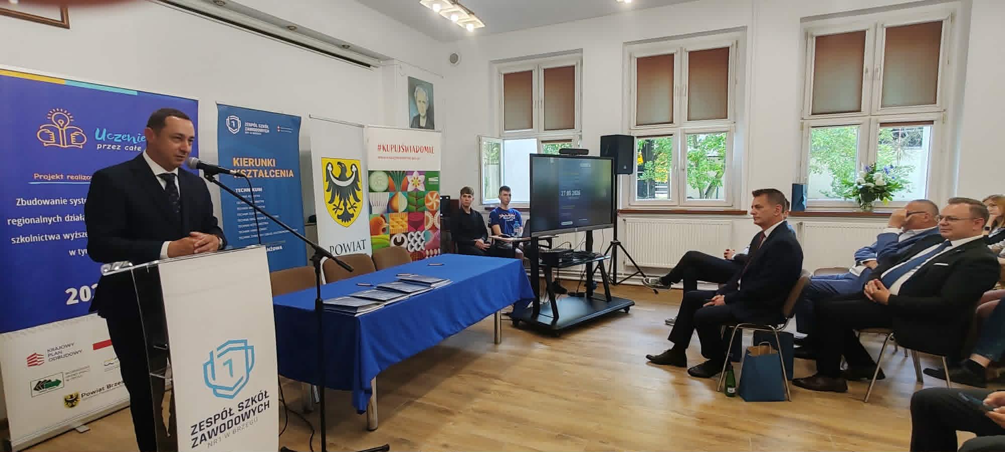 Mężczyzna w garniturze przemawia na podium w jasnym pomieszczeniu z siedzącymi uczestnikami. Za nim widoczne są banery, plakaty i ekran. Stoły z dokumentami znajdują się z przodu, a światło słoneczne wpada przez trzy wysokie okna w tle.