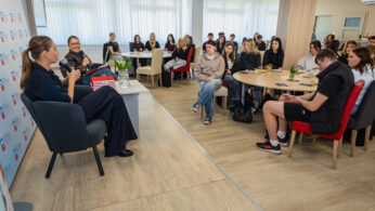 Kobieta mówi do mikrofonu podczas panelu dyskusyjnego w jasnym pomieszczeniu. Członkowie publiczności, głównie młodzi dorośli, słuchają uważnie, siedząc przy okrągłych stołach. Atmosfera jest swobodna, z notatnikami, laptopami i kwiatami na stołach.