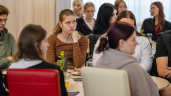 Grupa młodych ludzi siedzi wokół stołów, słuchając uważnie. Kobieta w brązowo-pomarańczowej koszuli w paski siedzi na pierwszym planie, opierając brodę na dłoni. Tło przedstawia jasny pokój z kilkoma osobami, które wydają się skupione i uważne.