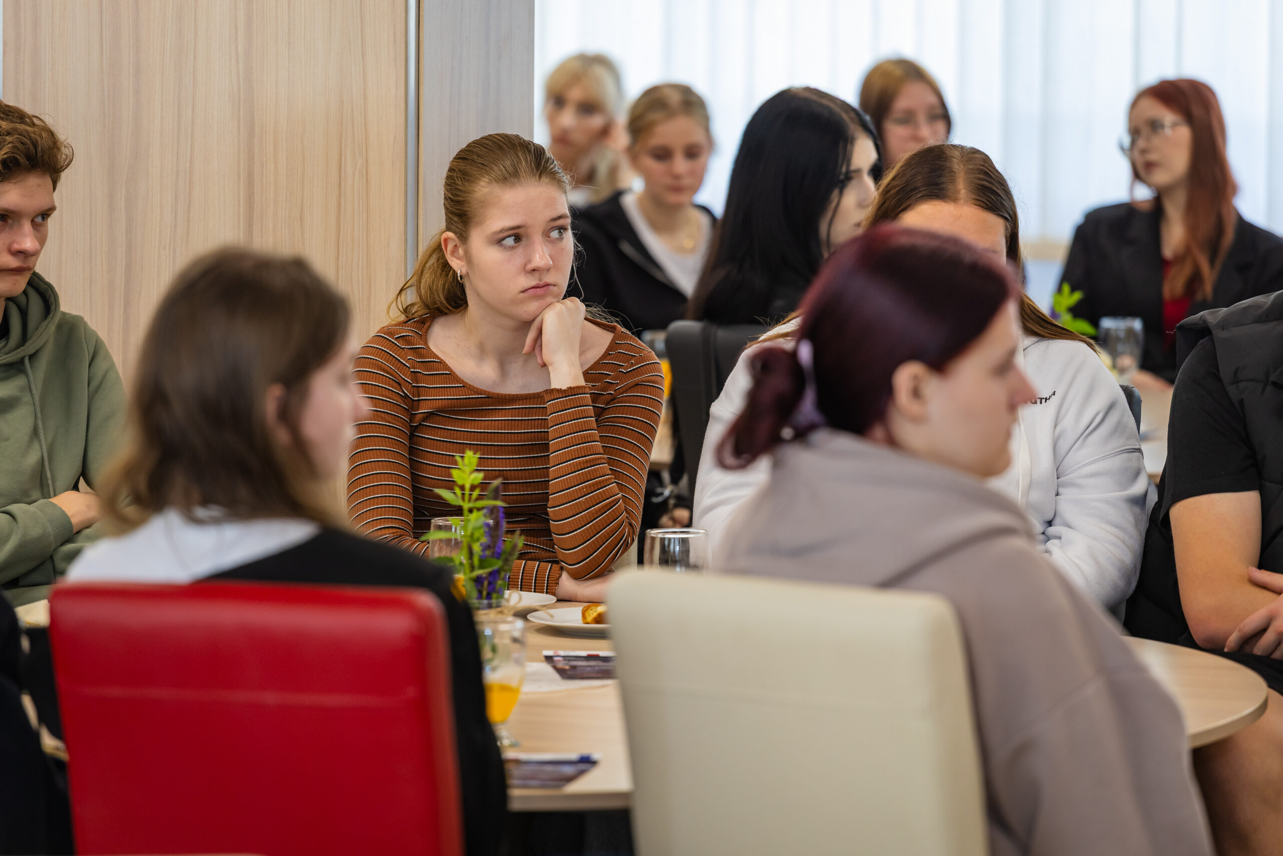Grupa młodych ludzi siedzi wokół stołów, słuchając uważnie. Kobieta w brązowo-pomarańczowej koszuli w paski siedzi na pierwszym planie, opierając brodę na dłoni. Tło przedstawia jasny pokój z kilkoma osobami, które wydają się skupione i uważne.