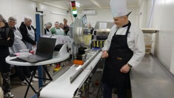 Kobieta w białej bluzie kucharskiej i ciemnych spodniach stoi po prawej stronie kadru, gestykulując dłońmi podczas mówienia do grupy młodzieży. Młodzież, ubrana w foliowe czepki i fartuchy ochronne, stoi w rzędzie po lewej stronie, słuchając uważnie. Na metalowym blacie w pobliżu kobiety leżą produkty spożywcze, w tym puszki z pomidorami i warzywa. W tle widać fragmenty profesjonalnej kuchni przemysłowej z metalowymi szafkami i piecem.