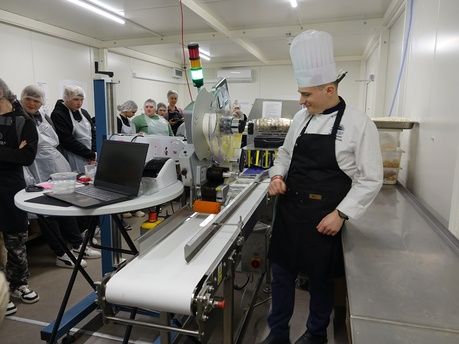 Kobieta w białej bluzie kucharskiej i ciemnych spodniach stoi po prawej stronie kadru, gestykulując dłońmi podczas mówienia do grupy młodzieży. Młodzież, ubrana w foliowe czepki i fartuchy ochronne, stoi w rzędzie po lewej stronie, słuchając uważnie. Na metalowym blacie w pobliżu kobiety leżą produkty spożywcze, w tym puszki z pomidorami i warzywa. W tle widać fragmenty profesjonalnej kuchni przemysłowej z metalowymi szafkami i piecem.