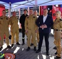 Siedem osób stoi w grupie pod błękitnym namiotem na zewnątrz na brukowanym placu. Pięć osób po lewej i jedna po prawej to strażacy w jasnobrązowych mundurach ochronnych. Między nimi stoi mężczyzna w ciemnym garniturze z rękami splecionymi przed sobą. W tle, za namiotem, widać część czerwonego wozu strażackiego.