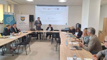 Szerokie ujęcie sali konferencyjnej podczas oficjalnej prezentacji prowadzonej przez mężczyznę w garniturze. Uczestnicy, zróżnicowani pod względem wieku, siedzą przy stołach w układzie litery U, mając przed sobą materiały szkoleniowe i napoje. Pomieszczenie jest nowoczesne, z kasetonowym sufitem i dużymi oknami zapewniającymi naturalne światło.
