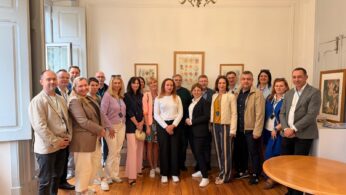 Oficjalne zdjęcie grupowe kilkunastu elegancko ubranych osób, pozujących w jasnym pomieszczeniu o klasycznym wystroju. Grupa stoi na drewnianym parkiecie pod ozdobnym żyrandolem, a niektórzy z nich mają na szyjach niebieskie smycze z identyfikatorami. Na białej ścianie w tle wiszą trzy symetrycznie rozmieszczone, oprawione obrazy.