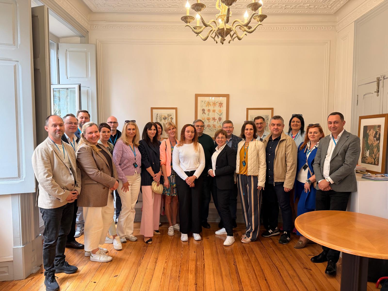 Oficjalne zdjęcie grupowe kilkunastu elegancko ubranych osób, pozujących w jasnym pomieszczeniu o klasycznym wystroju. Grupa stoi na drewnianym parkiecie pod ozdobnym żyrandolem, a niektórzy z nich mają na szyjach niebieskie smycze z identyfikatorami. Na białej ścianie w tle wiszą trzy symetrycznie rozmieszczone, oprawione obrazy.