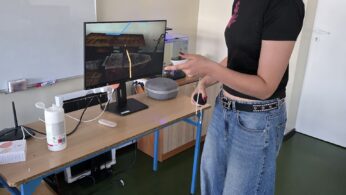Osoba nosząca biały zestaw VR i trzymająca kontrolery stoi w pobliżu biurka z monitorem wyświetlającym wirtualną scenę. Na biurku znajduje się szary głośnik, butelka ze środkiem odkażającym i komputer. Osoba ma na sobie czarną koszulkę i szerokie niebieskie dżinsy.