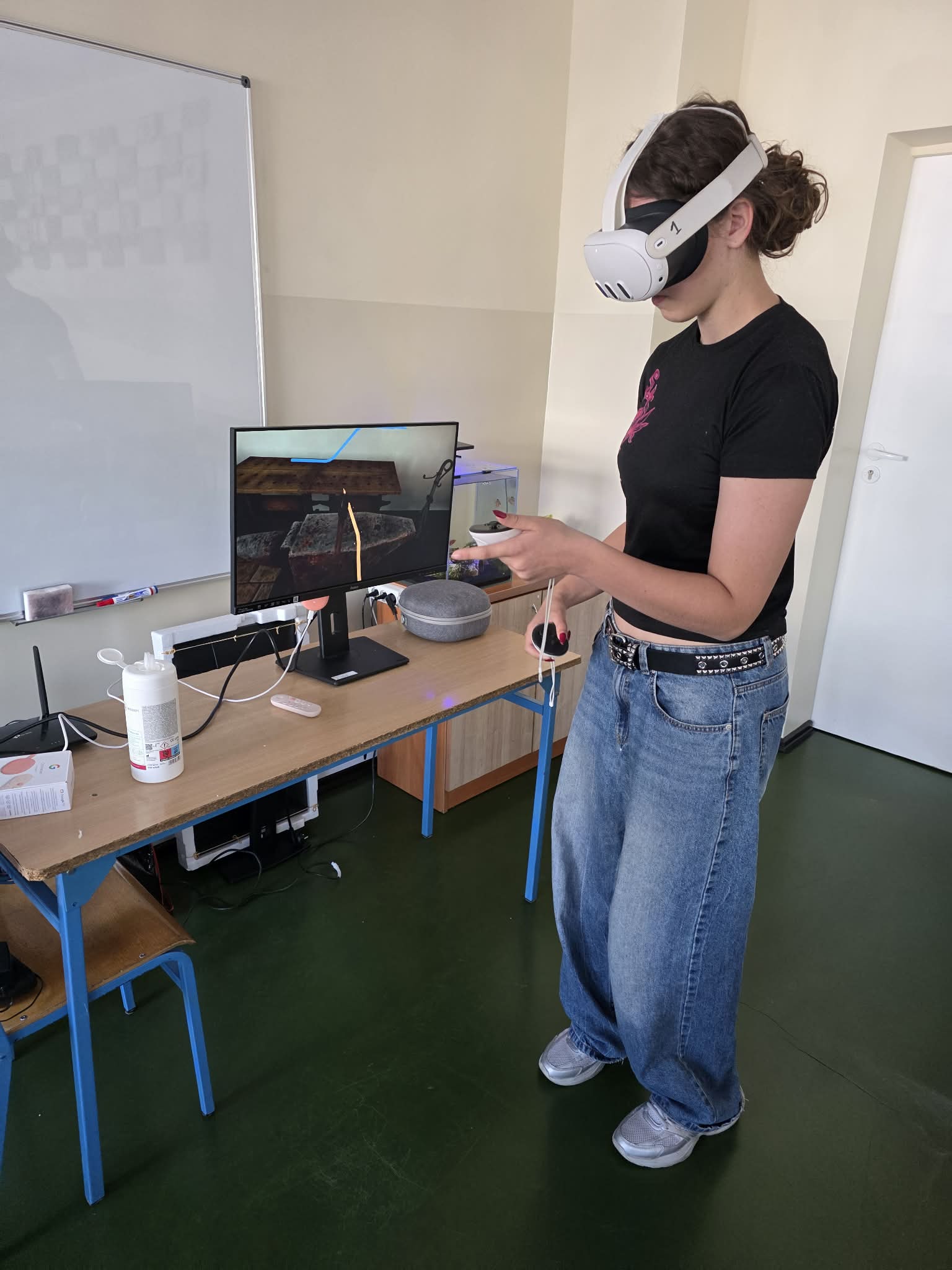 Osoba nosząca biały zestaw VR i trzymająca kontrolery stoi w pobliżu biurka z monitorem wyświetlającym wirtualną scenę. Na biurku znajduje się szary głośnik, butelka ze środkiem odkażającym i komputer. Osoba ma na sobie czarną koszulkę i szerokie niebieskie dżinsy.