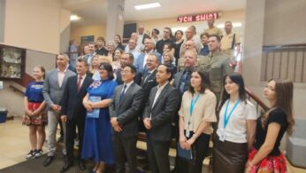 Duża grupa mężczyzn i kobiet, niektórzy w garniturach i mundurach wojskowych, inni w strojach codziennych lub tradycyjnych, pozują do oficjalnego zdjęcia w pomieszczeniu u podstawy schodów. W tle widoczne są gabloty z trofeami i czerwony podświetlany znak.