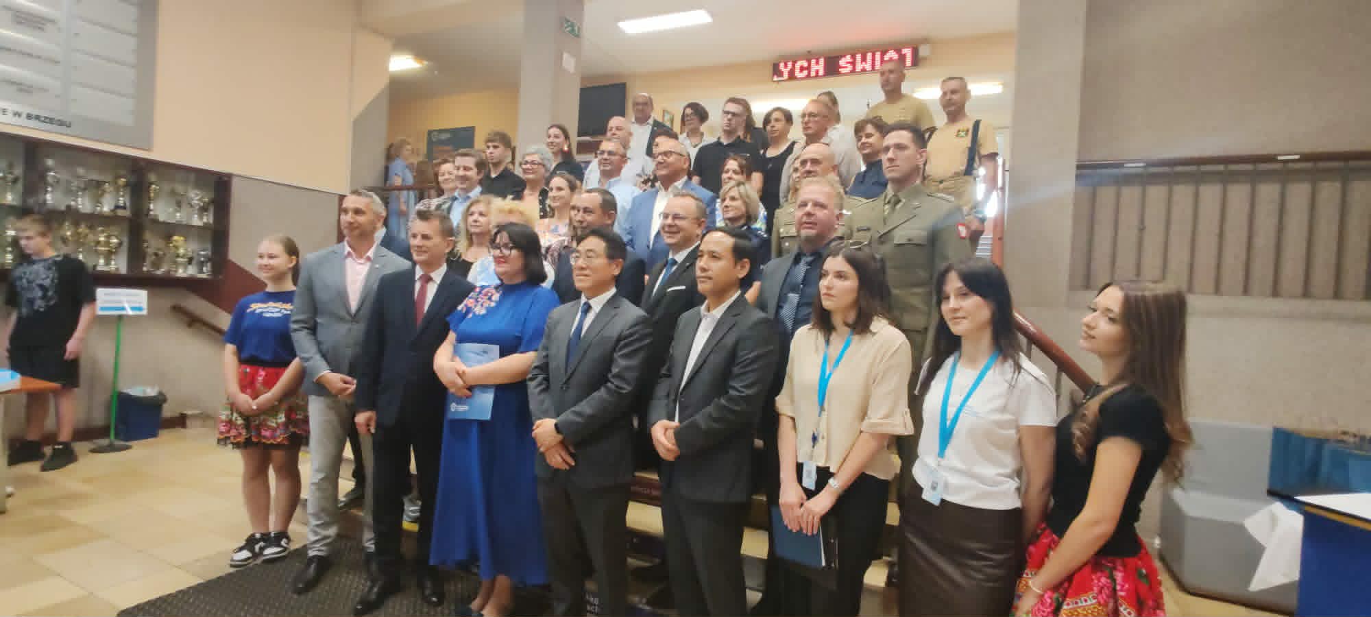 Duża grupa mężczyzn i kobiet, niektórzy w garniturach i mundurach wojskowych, inni w strojach codziennych lub tradycyjnych, pozują do oficjalnego zdjęcia w pomieszczeniu u podstawy schodów. W tle widoczne są gabloty z trofeami i czerwony podświetlany znak.