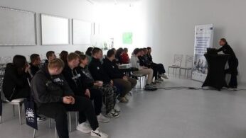 Liczna grupa osób, głównie młodzieży, pozuje do wspólnego zdjęcia na zewnątrz budynku. Stoją przed jasną elewacją, na której widnieje duży, niebieski napis „OPOLANKA”. Po lewej stronie ustawiony jest rollup z logotypami i tekstem dotyczącym wspierania kształcenia zawodowego. Niektóre osoby siedzą w kucki w pierwszym rzędzie, a jedna kobieta stoi z psem na rękach.