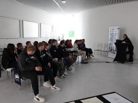 Liczna grupa osób, głównie młodzieży, pozuje do wspólnego zdjęcia na zewnątrz budynku. Stoją przed jasną elewacją, na której widnieje duży, niebieski napis „OPOLANKA”. Po lewej stronie ustawiony jest rollup z logotypami i tekstem dotyczącym wspierania kształcenia zawodowego. Niektóre osoby siedzą w kucki w pierwszym rzędzie, a jedna kobieta stoi z psem na rękach.