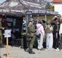 Grupa młodych ludzi w cywilnym ubraniu stoi obok wojskowego namiotu promocyjnego na świeżym powietrzu. Namiot ma ciemny wzór moro i napisy w języku polskim, w tym 