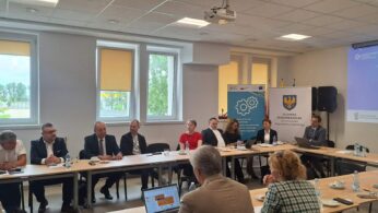 Zdjęcie dokumentuje przebieg konferencji, koncentrując się na rzędzie siedzących osób, które uważnie słuchają prelegenta. Na ekranie w tle wyświetlane są informacje o Zintegrowanym Systemie Kwalifikacji oraz logotypy Unii Europejskiej. Wnętrze jest minimalistyczne, a profesjonalny charakter spotkania podkreślają stroje biznesowe uczestników.
