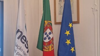 Trzy flagi stojące w pionowych stojakach wewnątrz eleganckiego pomieszczenia z jasnymi ścianami. Od lewej strony widoczna jest biała flaga z logotypem i napisem, w środku flaga Portugalii, a po prawej stronie niebieska flaga Unii Europejskiej z żółtymi gwiazdami. W tle, na ścianie z subtelnymi sztukateriami, wisi mały obraz w drewnianej ramie.