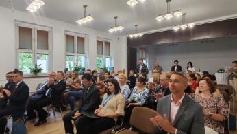 Grupa osób siedzi na brązowych krzesłach w dobrze oświetlonym pomieszczeniu z dużymi oknami i białymi ścianami. Większość z nich to osoby dorosłe, ubrane w stroje biznesowe lub eleganckie stroje codzienne, które wydają się uważnie obserwować lub słuchać czegoś poza kamerą.