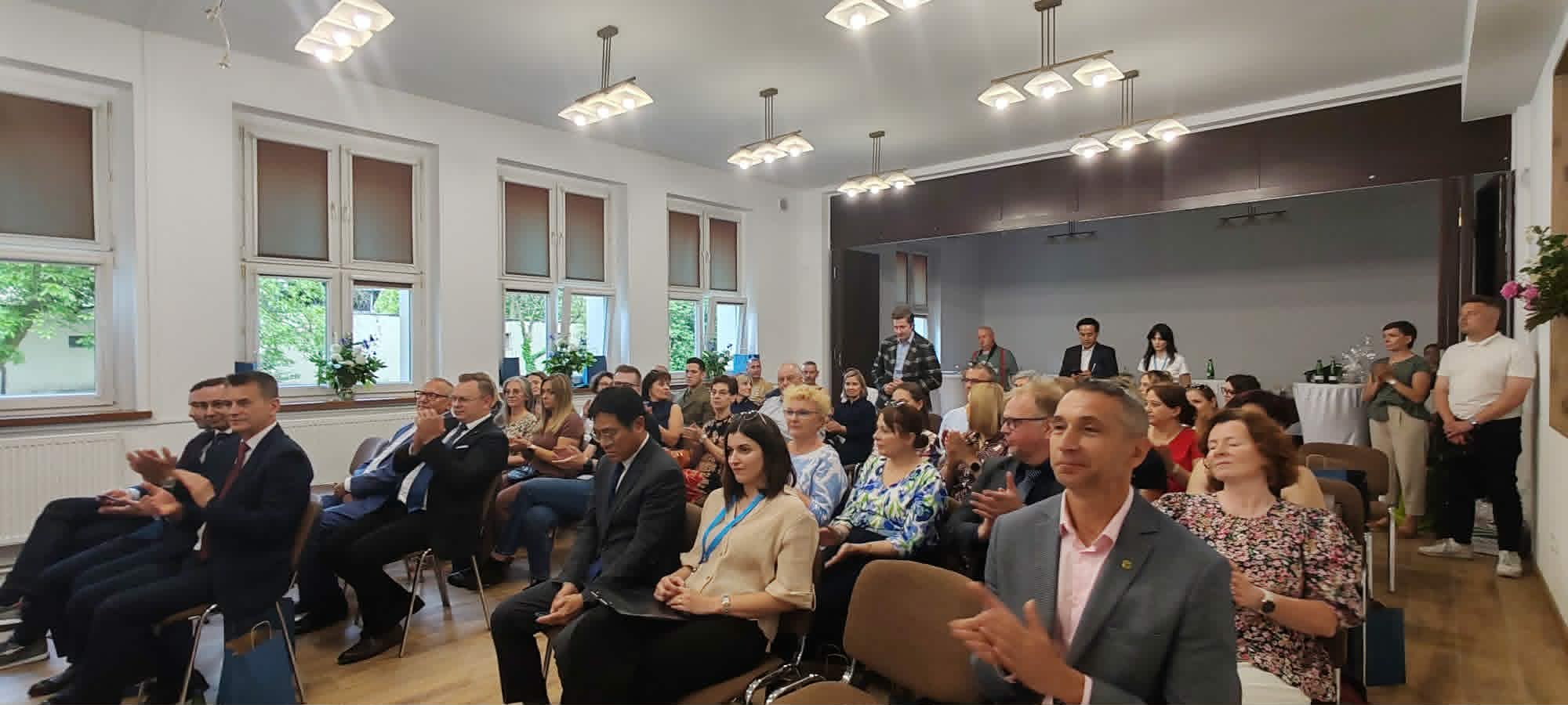Grupa osób siedzi na brązowych krzesłach w dobrze oświetlonym pomieszczeniu z dużymi oknami i białymi ścianami. Większość z nich to osoby dorosłe, ubrane w stroje biznesowe lub eleganckie stroje codzienne, które wydają się uważnie obserwować lub słuchać czegoś poza kamerą.