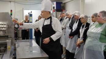 Dwie kobiety i mężczyzna siedzą przy długim, jasnym stole nakrytym czarnym obrusem, na którym stoją małe doniczki z zielonymi roślinami i filiżanki. Kobieta po lewej stronie kadru patrzy przed siebie z uśmiechem, słuchając kogoś poza kadrem. Po drugiej stronie stołu siedzi grupa młodzieży, skierowana w tym samym kierunku. W tle widać rollup z logotypami i tekstem dotyczącym wspierania kształcenia zawodowego.