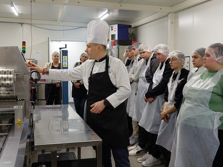 Dwie kobiety i mężczyzna siedzą przy długim, jasnym stole nakrytym czarnym obrusem, na którym stoją małe doniczki z zielonymi roślinami i filiżanki. Kobieta po lewej stronie kadru patrzy przed siebie z uśmiechem, słuchając kogoś poza kadrem. Po drugiej stronie stołu siedzi grupa młodzieży, skierowana w tym samym kierunku. W tle widać rollup z logotypami i tekstem dotyczącym wspierania kształcenia zawodowego.