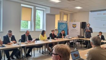 Fotografia przedstawia moment prelekcji w sali obrad, gdzie mężczyzna w okularach i garniturze tłumaczy założenia projektu widocznego na slajdzie. Uczestnicy są zwróceni w stronę mówcy, a na stołach przed nimi znajdują się laptopy, notatniki oraz poczęstunek. Po lewej stronie widać duże okna z widokiem na zieleń, co rozjaśnia całe pomieszczenie.