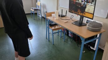 Dziecko noszące zestaw VR stoi w klasie, naprzeciwko biurka z monitorem wyświetlającym scenę kuchenną. Inna osoba wskazuje na ekran. Na biurku znajduje się klawiatura, mysz i urządzenia elektroniczne. Klasa ma zielone podłogi i tablice.