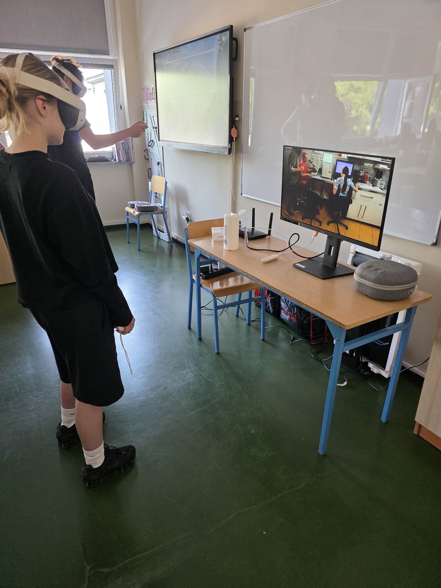 Dziecko noszące zestaw VR stoi w klasie, naprzeciwko biurka z monitorem wyświetlającym scenę kuchenną. Inna osoba wskazuje na ekran. Na biurku znajduje się klawiatura, mysz i urządzenia elektroniczne. Klasa ma zielone podłogi i tablice.