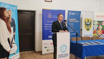 Mężczyzna w garniturze przemawia na mównicy z mikrofonami, obok niebieskiego stołu z dokumentami. Młoda kobieta stoi po lewej stronie banerów. Kolorowe emblematy hrabstwa i szkoły oraz plakaty są widoczne na ścianie za nimi.